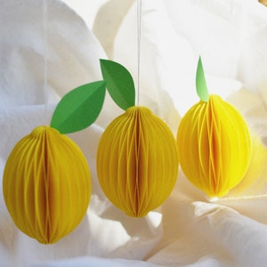 May include: Three yellow paper lemon ornaments with green paper leaves hanging from white string on a white fabric background.