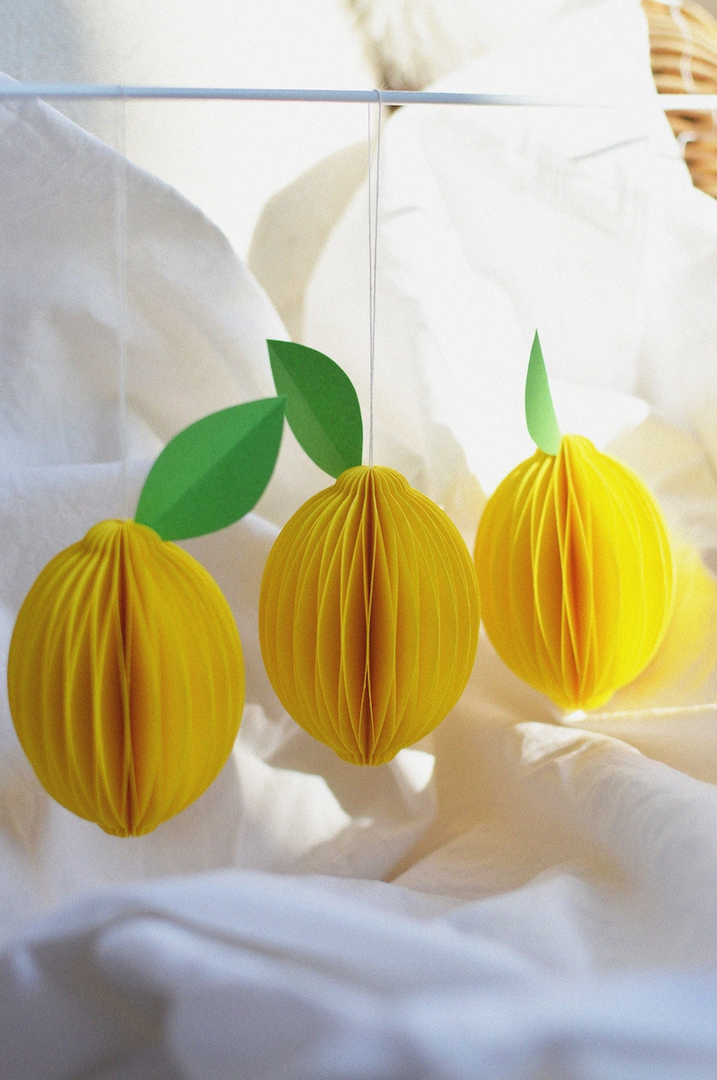 May include: Three yellow paper lemon ornaments with green paper leaves hanging from white string on a white fabric background.