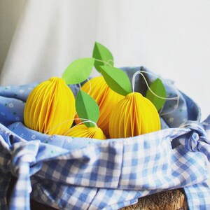 May include: Four yellow paper lemons with green paper leaves in a blue and white gingham fabric bag on a wooden surface.