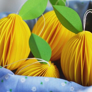 May include: Four yellow paper sphere ornaments with green paper leaves attached. The ornaments are sitting on a blue fabric with a white floral pattern.