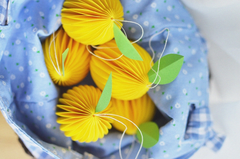 May include: A blue fabric bag with white polka dots holds five yellow paper honeycomb balls with green paper leaves attached.