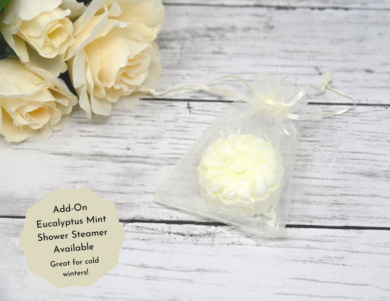 May include: A white shower steamer in a clear organza bag. The steamer is shaped like a flower and has a light green tint. The text "Add-On Eucalyptus Mint Shower Steamer Available Great for cold winters!" is written on a white background.