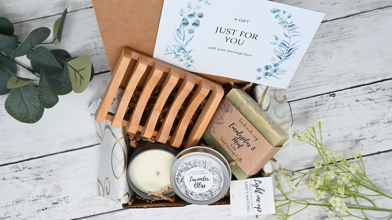 May include: A gift box filled with self-care items, including a candle, soap, and a wooden soap dish. The box is decorated with a card that says "A Gift Just For You" and a small bag of matches that says "Light me up".