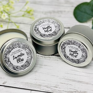 May include: Three silver candle tins with black and white labels. The labels feature a floral design and the text "Hand Poured Soy Wax Candles". The candles are labeled "Lavender Bliss", "Vanilla Velvet", and "Vanilla Velvet".