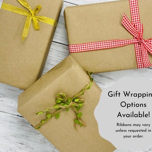 May include: Three brown paper gift boxes with different ribbon styles. The boxes are tied with a gold ribbon, a red and white gingham ribbon, and a brown twine ribbon with green leaves. The text on the image reads "Gift Wrapping Options Available! Ribbons may vary unless requested in your order."