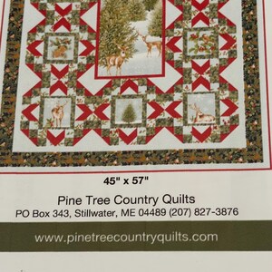 May include: A quilt featuring a winter forest scene with deer, pine trees, and holly berries in red, green, and white. The pattern includes geometric blocks and a decorative border. Text reads 45" x 57", Pine Tree Country Quilts, Stillwater, ME.