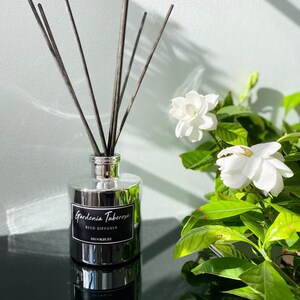 May include: A silver bottle with a black label that reads "Gardenia Tuberose Reed Diffuser" and "BROOKBURY". The bottle is filled with black reed sticks and is sitting on a black surface. There are white flowers in the background.