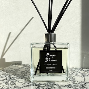 May include: A clear glass bottle with a black label that reads "Orange Blossom Reed Diffuser Brookbury 6 fl oz". The bottle is filled with a clear liquid and has black reeds sticking out of the top.