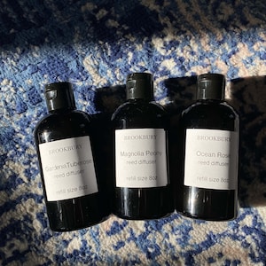 May include: Three black glass bottles with white labels. The labels read "BROOKBURY" and list different reed diffuser scents: "Gardenia Tuberose", "Magnolia Peony", and "Ocean Rose". Each bottle is labeled with "refill size 8oz".