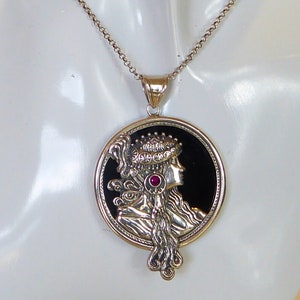Könnte beinhalten: Eine silberne Halskette mit einem runden Anhänger, der ein schwarzes Kamee mit dem Profil einer Frau mit einem rosa Edelsteinakzent zeigt. Der Anhänger ist mit aufwendigen Silberarbeiten verziert.
