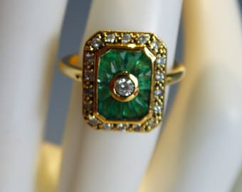 Può includere: Un anello d'oro con un design rettangolare di smeraldi e diamanti. Lo smeraldo è circondato da un bordo di piccoli diamanti. L'anello è posto su una superficie bianca.