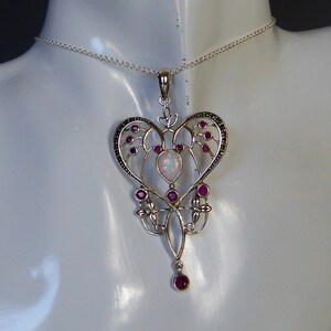 Puede incluir: Un collar de plata con un colgante en forma de corazón que presenta una ópalo blanca y piedras preciosas rosas. El colgante está detallado con un intrincado trabajo de filigrana y cuelga de una cadena de plata.