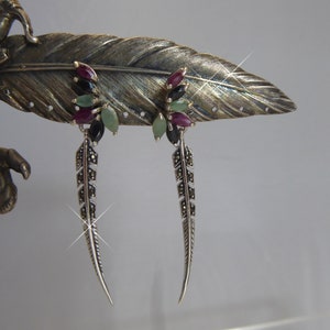 May include: Silver feather earrings with ruby, emerald, and onyx accents. The earrings are displayed on a decorative silver feather. The earrings have a long, slender design with a detailed pattern.