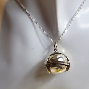 Puede incluir: Collar con colgante de medallón plateado, redondo y articulado. El colgante presenta un diseño detallado y texturizado, suspendido de una fina cadena. El medallón se exhibe sobre un maniquí blanco.