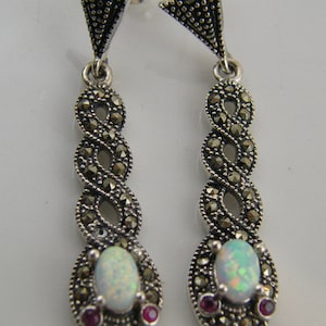 May include: A pair of silver dangle earrings featuring a white opal gemstone and two ruby accents. The earrings are crafted with a twisted design of black marcasite.