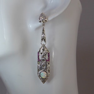 May include: A pair of silver drop earrings featuring a teardrop-shaped opal stone and a geometric design accented with small red stones.