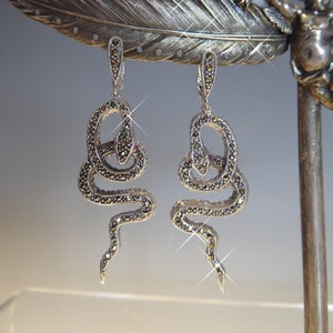 Peut inclure: Boucles d'oreilles serpent en argent avec détails en marcassite et une petite pierre rouge. Les boucles d'oreilles sont présentées sur une plume décorative en argent. Le motif serpent est populaire en bijouterie.