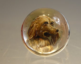 May include: A silver ring with a clear glass cabochon featuring a hand-painted portrait of a brown dog. The dog's head is turned to the side, and its eyes are visible.