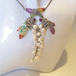 May include: A necklace featuring two metallic insect pendants with multicolored enamel wings in shades of green, purple, and red. Each insect has a small dark red stone eye and holds a cluster of white oval pearls hanging from a metal chain.