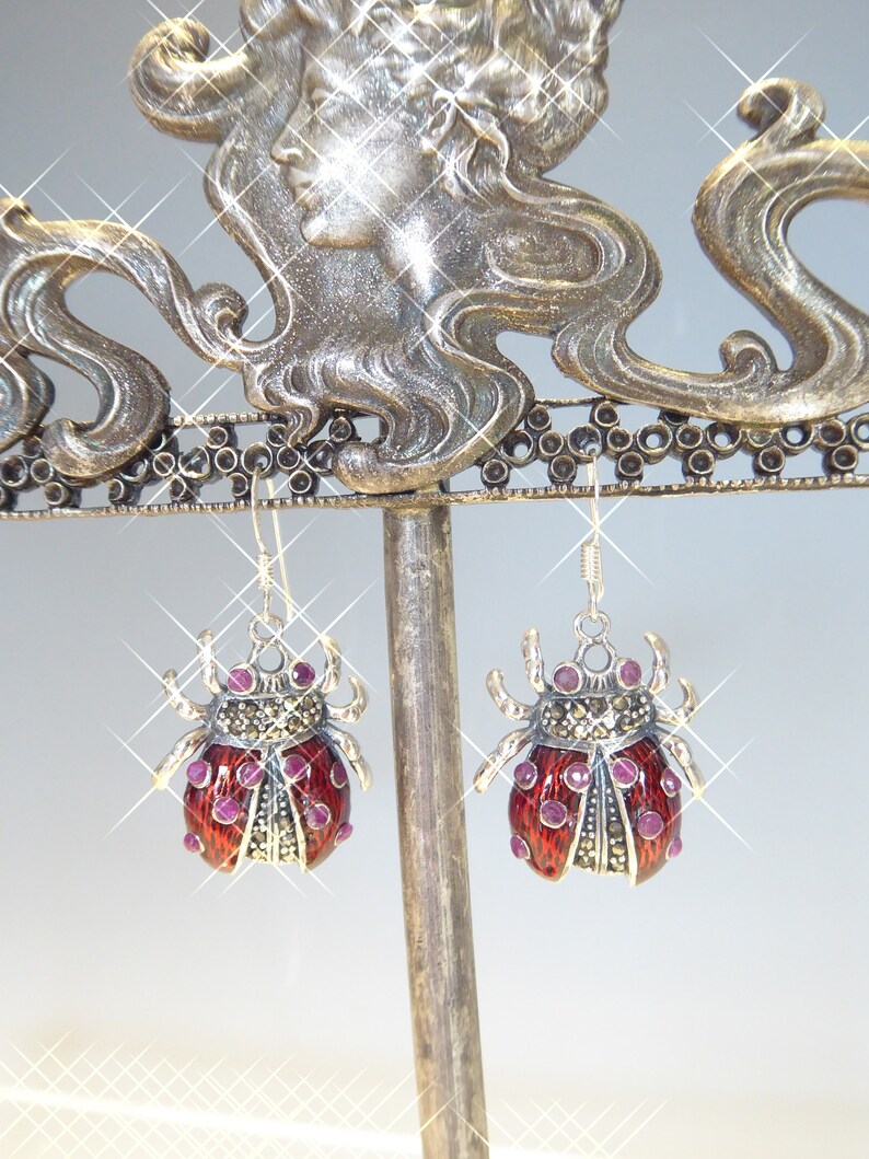 May include: A pair of silver earrings shaped like ladybugs with red enamel and pink gemstone accents.