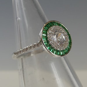 May include: A silver ring featuring a large round clear stone encircled by a halo of smaller clear stones and a border of green stones.