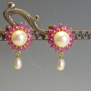 May include: A pair of gold earrings with pink gemstone accents and a pearl drop. The earrings feature a cluster of pink gemstones surrounding a pearl.