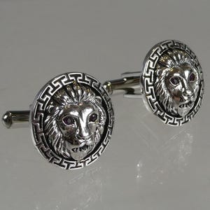 May include: Silver cufflinks with a lion head design. The lion's eyes are set with pink gemstones. The cufflinks are surrounded by a decorative border.