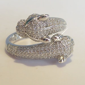 Könnte beinhalten: Ein silberner Ring mit zwei Tierköpfen auf jeder Seite. Der Ring ist mit kleinen, klaren Steinen besetzt.