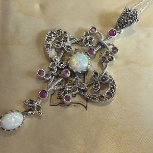 Puede incluir: Un collar con un colgante de plata adornado con una piedra de ópalo blanco y un diseño delicado de marcasita y piedras rosas. El colgante está colgado de una cadena de plata.