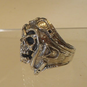 Peut inclure: Une bague argentée avec un motif de crâne. La bague présente un visage de crâne détaillé avec des détails métalliques complexes autour de la tête et des côtés. La bague a une esthétique gothique unique.