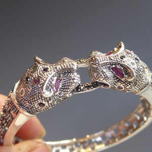 May include: Silver bracelet with two jaguar head motifs. The heads are embellished with small, colourful gemstones, including red, green, and black. The bracelet is hinged. The design is intricate and detailed.