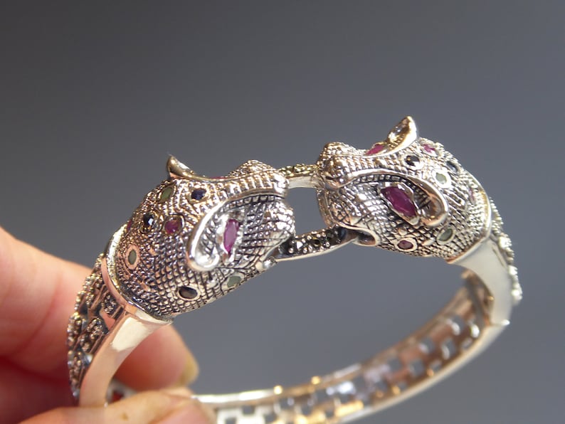 May include: Silver bracelet with two jaguar head motifs. The heads are embellished with small, colourful gemstones, including red, green, and black. The bracelet is hinged. The design is intricate and detailed.