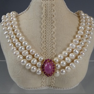 May include: A three-strand necklace featuring white freshwater pearls arranged in multiple rows. The center of the necklace is adorned with a large, oval-shaped pink cabochon stone surrounded by a cluster of small, vibrant red faceted gemstones.