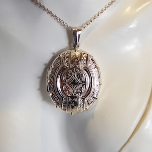 Puede incluir: Un medallón ovalado de plata con un intrincado diseño floral en la parte delantera. El medallón está unido a una cadena de plata.