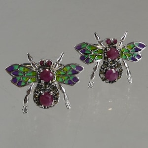 May include: A pair of silver earrings shaped like bees with green, purple, and pink enamel. The bees have ruby gemstones for eyes and black gemstones for the body.