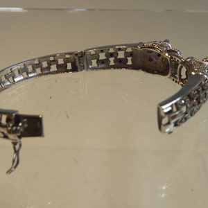 May include: Silver bracelet with an open design. The bracelet has a decorative clasp and is decorated with small, sparkling stones and a detailed animal head design. The bracelet is open, showing the clasp and the intricate details.