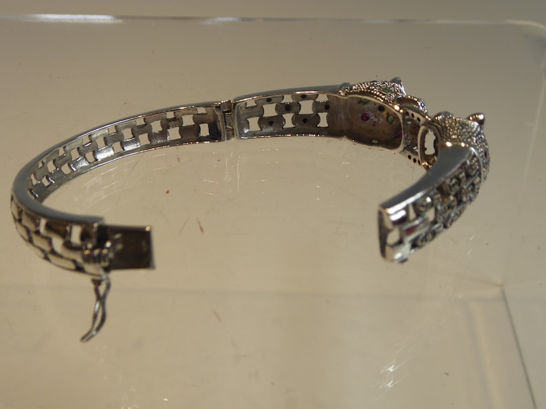 May include: Silver bracelet with an open design. The bracelet has a decorative clasp and is decorated with small, sparkling stones and a detailed animal head design. The bracelet is open, showing the clasp and the intricate details.