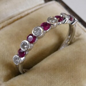 May include: A silver ring with alternating round gemstones. The ring features clear, sparkling stones and deep red rubies. The ring is set in a light-coloured jewellery box.