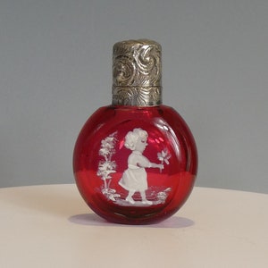 May include: A red glass bottle with a silver top and a white cameo of a girl holding a flower. The bottle is round and has a smooth surface.