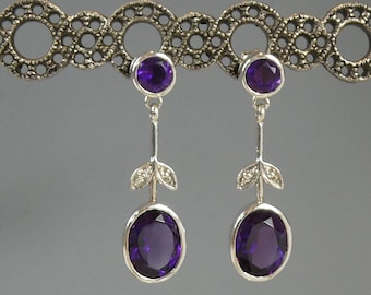 May include: A pair of silver drop earrings featuring oval and round purple gemstones. The earrings have a leaf design with small clear stones. The earrings are displayed on a decorative silver bar.