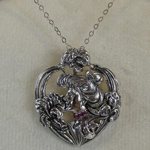 Könnte beinhalten: Silberfarbene Anhängerkette mit dem Profil einer Frau, umgeben von floralen und herzförmigen Designs. Der Hals der Frau ist mit kleinen roten Edelsteinen besetzt. Die Halskette hängt an einer filigranen Kette.