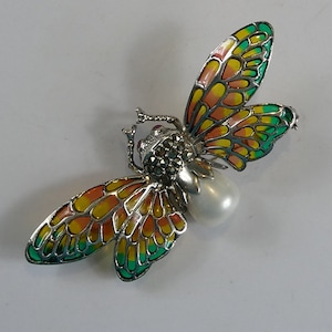 Könnte beinhalten: Eine silberne Brosche mit einer Perle und einem bunten Schmetterlingsdesign. Die Schmetterlingsflügel sind grün, gelb und orange.