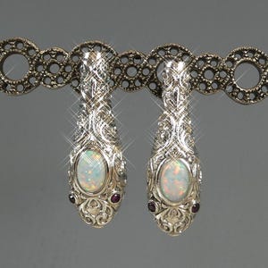May include: A pair of ornate silver earrings with an oval opal center stone and two small red gemstones. The earrings have a detailed, swirling design and are displayed on a decorative silver bar.