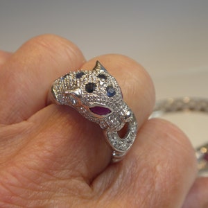 May include: A silver ring shaped like a panther's head, embellished with small, sparkling stones. The panther's eyes are blue, and the mouth features a red stone. A matching bracelet is visible in the background.