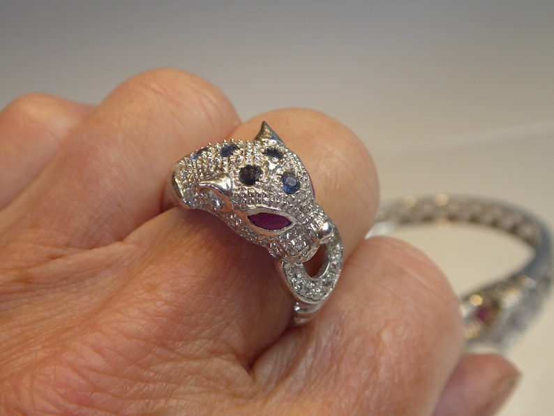 May include: A silver ring shaped like a panther's head, embellished with small, sparkling stones. The panther's eyes are blue, and the mouth features a red stone. A matching bracelet is visible in the background.