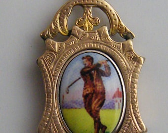 Può includere: Ciondolo dorato con un centro ovale in smalto raffigurante un golfista in azione. Il ciondolo ha una cornice ornata con un anello superiore decorativo. Il golfista è vestito di marrone ed è ambientato su un campo verde e un cielo azzurro.