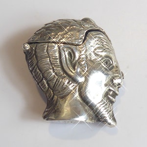 May include: A silver-colored, hinged box shaped like a man's head with a beard. The box has detailed facial features and hair, and the lid is slightly ajar. The surface of the box has a shiny, reflective quality.