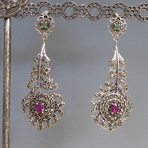 May include: A pair of silver earrings with a feather design. The earrings are adorned with marcasite stones and feature a pink gemstone in the center of each feather.