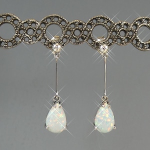 May include: A pair of drop earrings featuring pear-shaped white opals suspended from thin silver-toned metal bars. The earrings are attached to a decorative, dark silver-toned metal band with a repeating circular, filigree-style pattern.