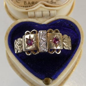 May include: A gold ring featuring a central vertical bar set with three clear stones, flanked by two round red stones, each encased in a star-shaped setting. The band displays intricate cut-out details and engraved patterns on the sides.
